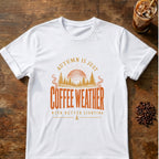 Vintage-style typography reading Autumn Is Just Coffee Weather With Better Lighting over a sunset, pine trees and steaming coffee swirls on a White T-shirt.