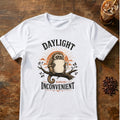 Sleepy owl monkey hugging a giant steaming mug on a branch in front of a rising sun with the words Daylight Remains Inconvenient on a White T-shirt.