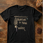 Tired bat hanging upside down from a branch holding a spilling coffee mug beside hanging letters reading Hanging in There. Barely on a Black T-shirt.