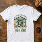 Content moose leaning out of a rustic cabin window holding a mug among autumn leaves with the words Outside Is Beautiful. I'll Be Inside on a White T-shirt.