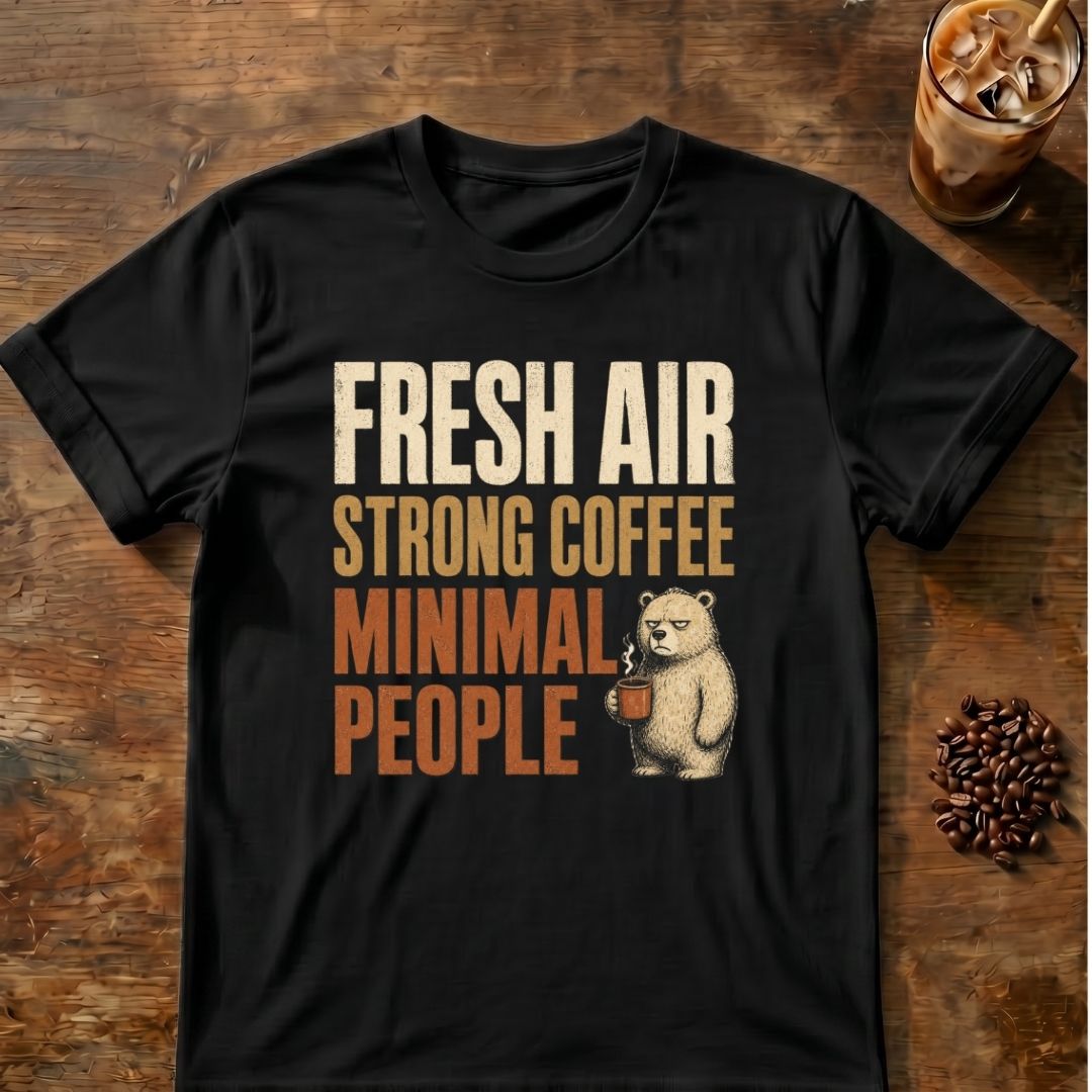 A black short-sleeve graphic T-shirt laid flat on a rustic wooden surface, with a printed slogan reading “FRESH AIR STRONG COFFEE MINIMAL PEOPLE” in cream and rust tones and a small bear holding a coffee cup. A glass of iced coffee and scattered coffee beans appear as styled props around the shirt.