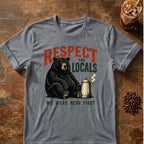 A heathered graphite-gray graphic t-shirt laid flat on a rustic wooden table, featuring a black bear sitting next to a coffee mug with the text 'RESPECT THE LOCALS WE WERE HERE FIRST'. Coffee beans and an iced coffee are styled around the shirt.