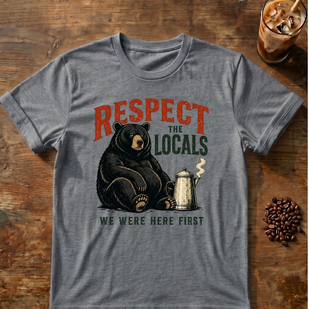 A heathered graphite-gray graphic t-shirt laid flat on a rustic wooden table, featuring a black bear sitting next to a coffee mug with the text 'RESPECT THE LOCALS WE WERE HERE FIRST'. Coffee beans and an iced coffee are styled around the shirt.