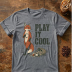 A graphite heather short-sleeve graphic T-shirt laid flat on a rustic wooden surface, featuring an illustration of a fox holding a mug next to the text 'PLAY IT COOL' and smaller text 'they know nothing.' Coffee-themed props, including an iced coffee glass and coffee beans, are placed around the shirt.