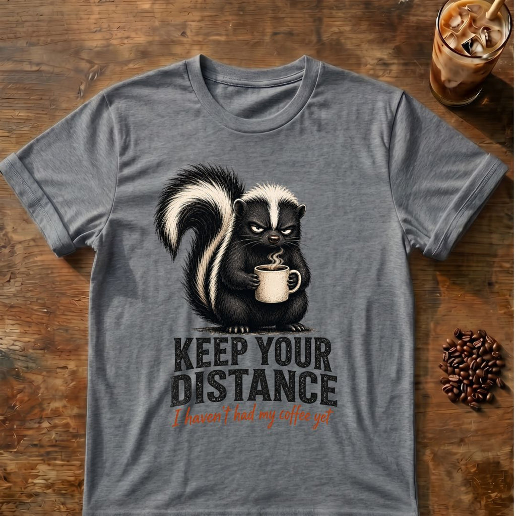 Grumpy skunk holding a steaming coffee mug above distressed text reading Keep Your Distance, with orange script saying I haven’t had my coffee yet. Graphite Heather
