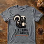 Grumpy skunk holding a steaming coffee mug above distressed text reading Keep Your Distance, with orange script saying I haven’t had my coffee yet. Graphite Heather