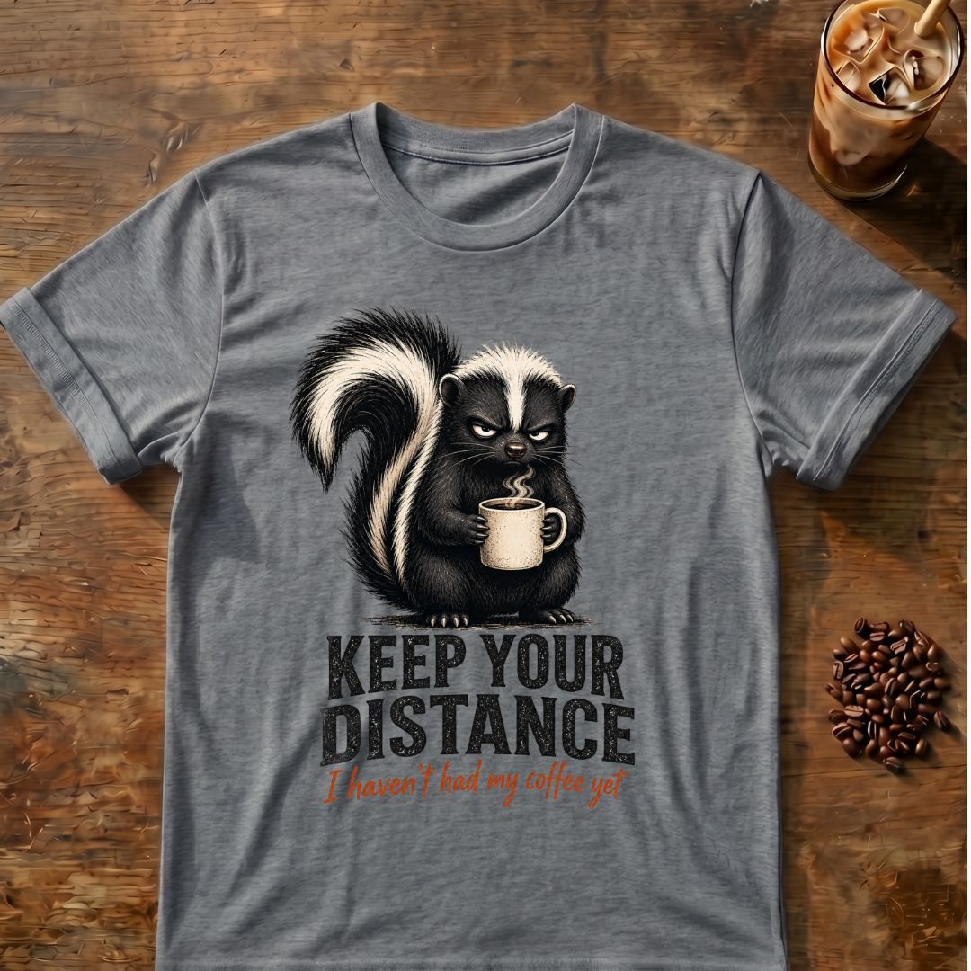 Grumpy skunk holding a steaming coffee mug above distressed text reading Keep Your Distance, with orange script saying I haven’t had my coffee yet. Graphite Heather