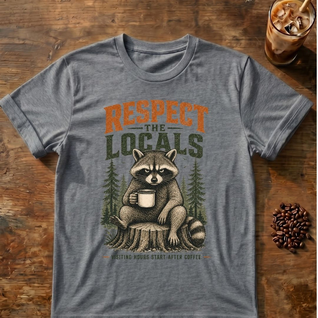 A heather gray graphic t-shirt featuring a raccoon sitting on a tree stump and holding a coffee mug, with the text 'RESPECT THE LOCALS' and 'COFFEE HUNTERS START AFTER COFFEE'. The t-shirt is displayed flat on a rustic wooden table with coffee beans and an iced coffee in the background.