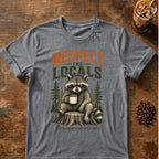 A heather gray graphic t-shirt featuring a raccoon sitting on a tree stump and holding a coffee mug, with the text 'RESPECT THE LOCALS' and 'COFFEE HUNTERS START AFTER COFFEE'. The t-shirt is displayed flat on a rustic wooden table with coffee beans and an iced coffee in the background.