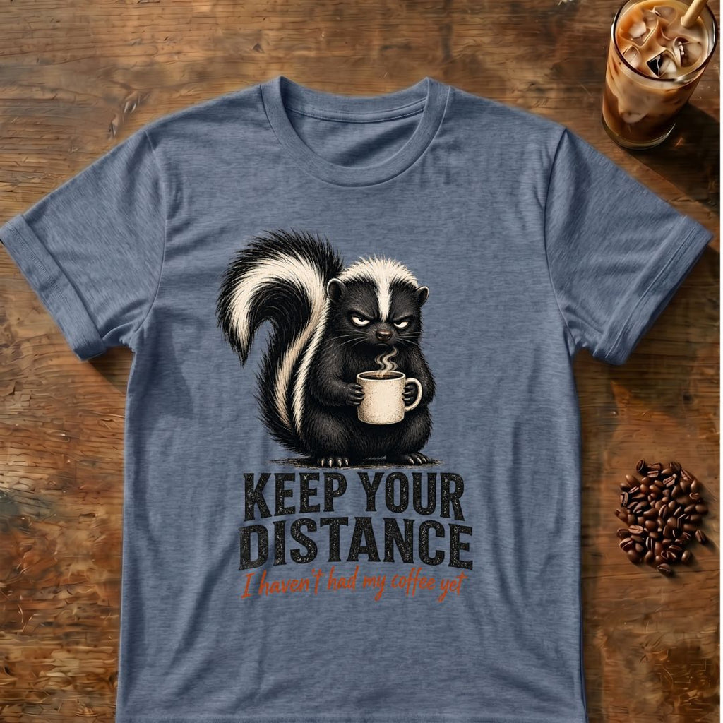 Grumpy skunk holding a steaming coffee mug above distressed text reading Keep Your Distance, with orange script saying I haven’t had my coffee yet. Indigo Heather
