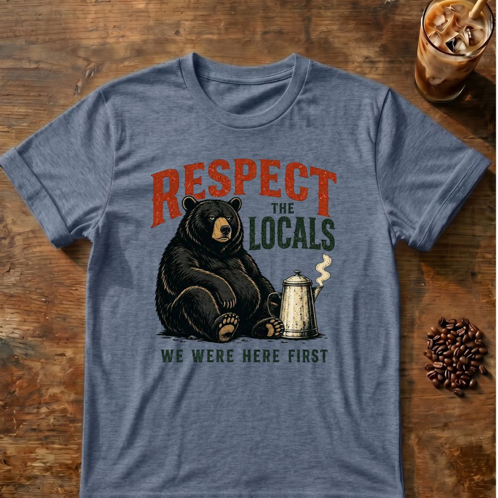 A heathered blue-gray graphic t-shirt laid flat on a rustic wooden table, featuring a black bear sitting next to a coffee mug with the text 'RESPECT THE LOCALS WE WERE HERE FIRST'. Coffee beans and an iced coffee are styled around the shirt.