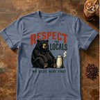 A heathered blue-gray graphic t-shirt laid flat on a rustic wooden table, featuring a black bear sitting next to a coffee mug with the text 'RESPECT THE LOCALS WE WERE HERE FIRST'. Coffee beans and an iced coffee are styled around the shirt.