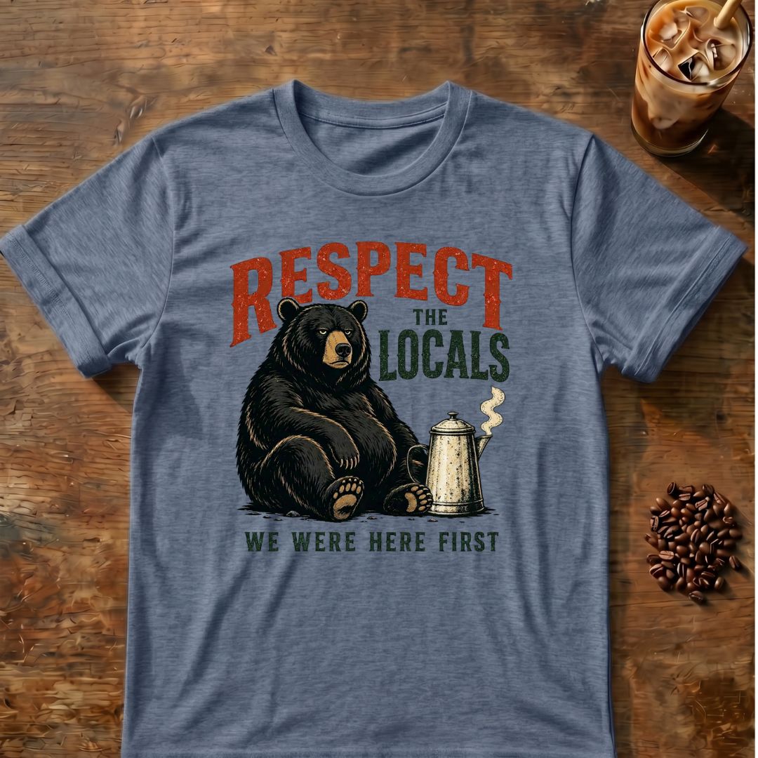 A heathered blue-gray graphic t-shirt laid flat on a rustic wooden table, featuring a black bear sitting next to a coffee mug with the text 'RESPECT THE LOCALS WE WERE HERE FIRST'. Coffee beans and an iced coffee are styled around the shirt.