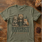An olive green graphic t-shirt laid flat on a rustic wooden table, featuring an illustration of a bear, owl, raccoon, and fox sharing coffee mugs with the text 'RESPECT THE LOCALS' and 'EAT LIFE COFFEE LIFE'. A glass of iced coffee and coffee beans are styled as props in the corners.
