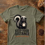 Grumpy skunk holding a steaming coffee mug above distressed text reading Keep Your Distance, with orange script saying I haven’t had my coffee yet. Heather Army Green
