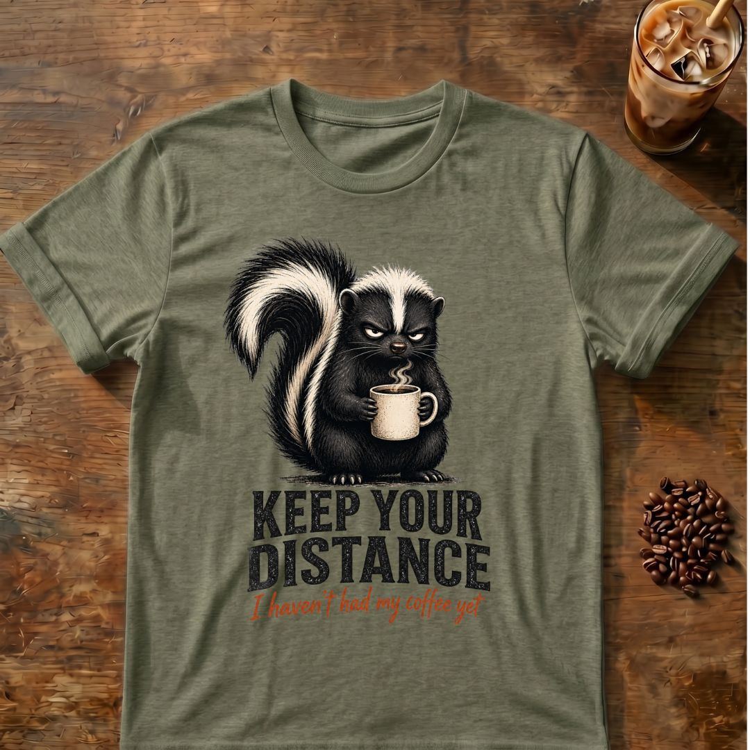 Grumpy skunk holding a steaming coffee mug above distressed text reading Keep Your Distance, with orange script saying I haven’t had my coffee yet. Heather Army Green