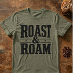 An olive green heather t-shirt with a distressed black graphic reading 'ROAST & ROAM' laid flat on a rustic wooden table, styled with coffee beans and an iced coffee in the background.