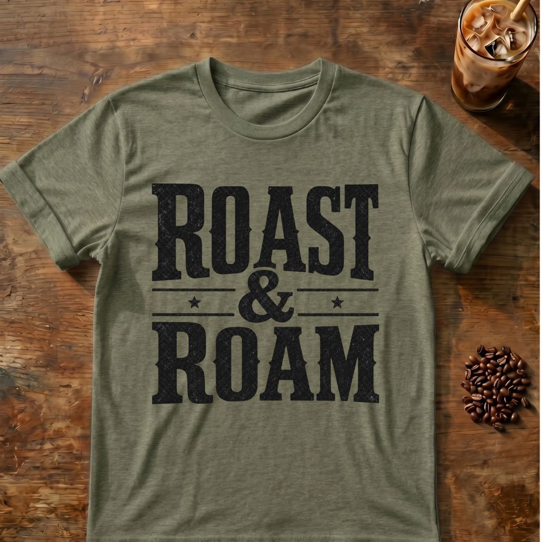 An olive green heather t-shirt with a distressed black graphic reading 'ROAST & ROAM' laid flat on a rustic wooden table, styled with coffee beans and an iced coffee in the background.