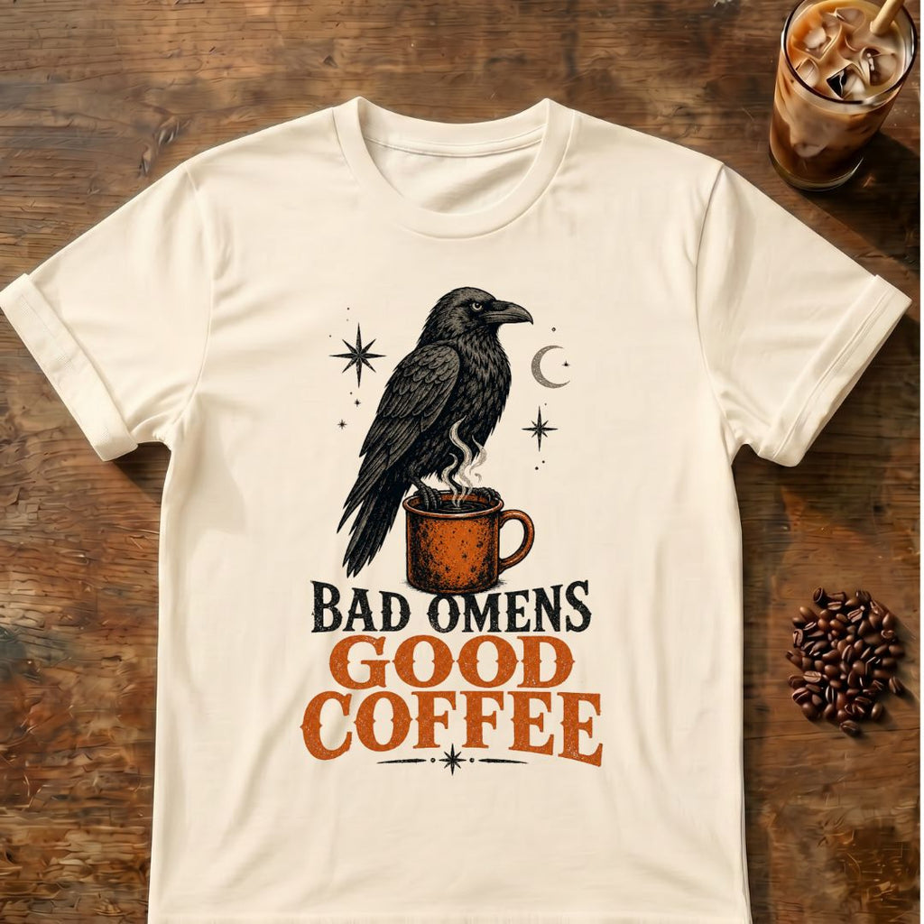 Black raven perched on a steaming orange coffee mug beneath a crescent moon and stars with the words Bad Omens, Good Coffee.