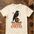 Black raven perched on a steaming orange coffee mug beneath a crescent moon and stars with the words Bad Omens, Good Coffee.