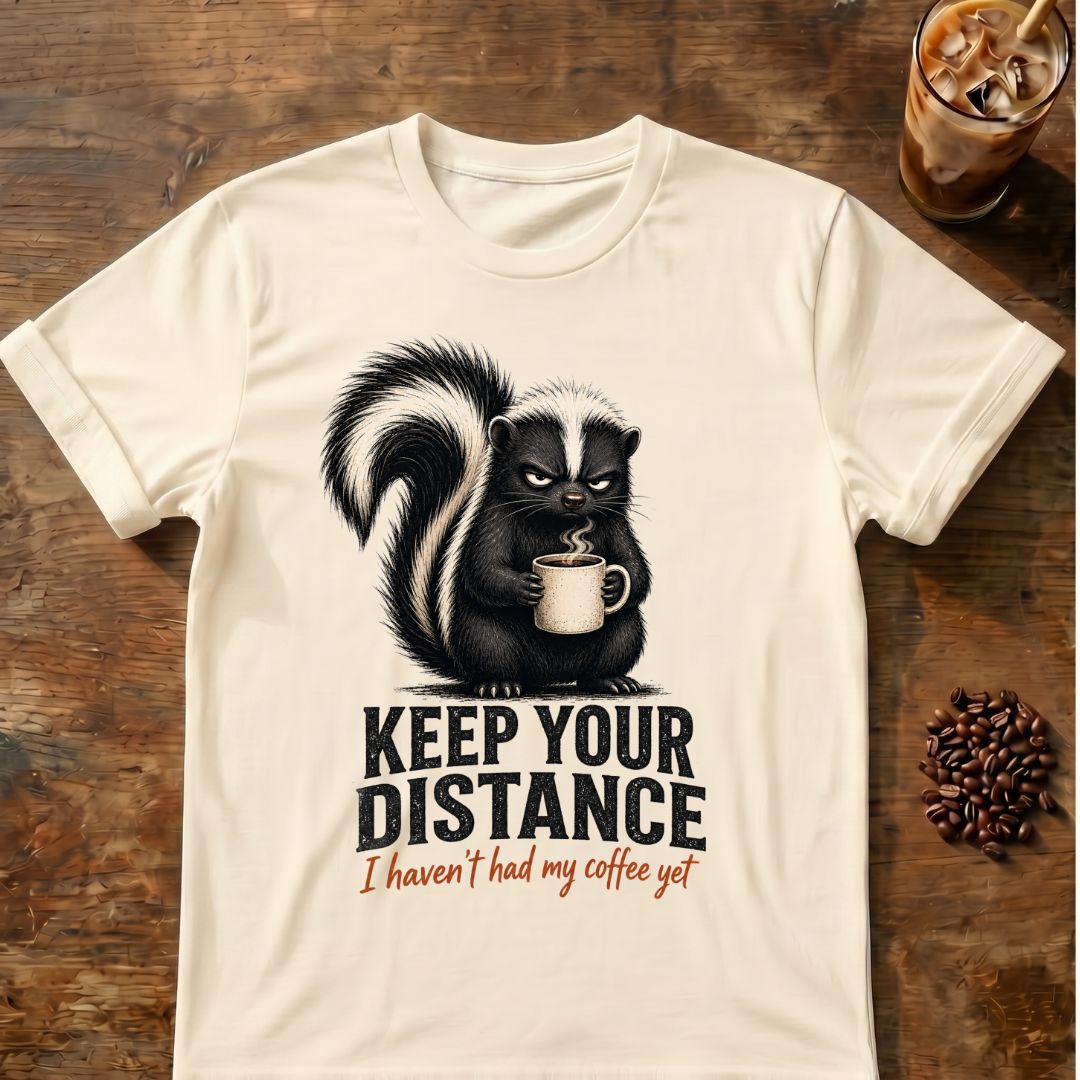 Grumpy skunk holding a steaming coffee mug above distressed text reading Keep Your Distance, with orange script saying I haven’t had my coffee yet. Natural