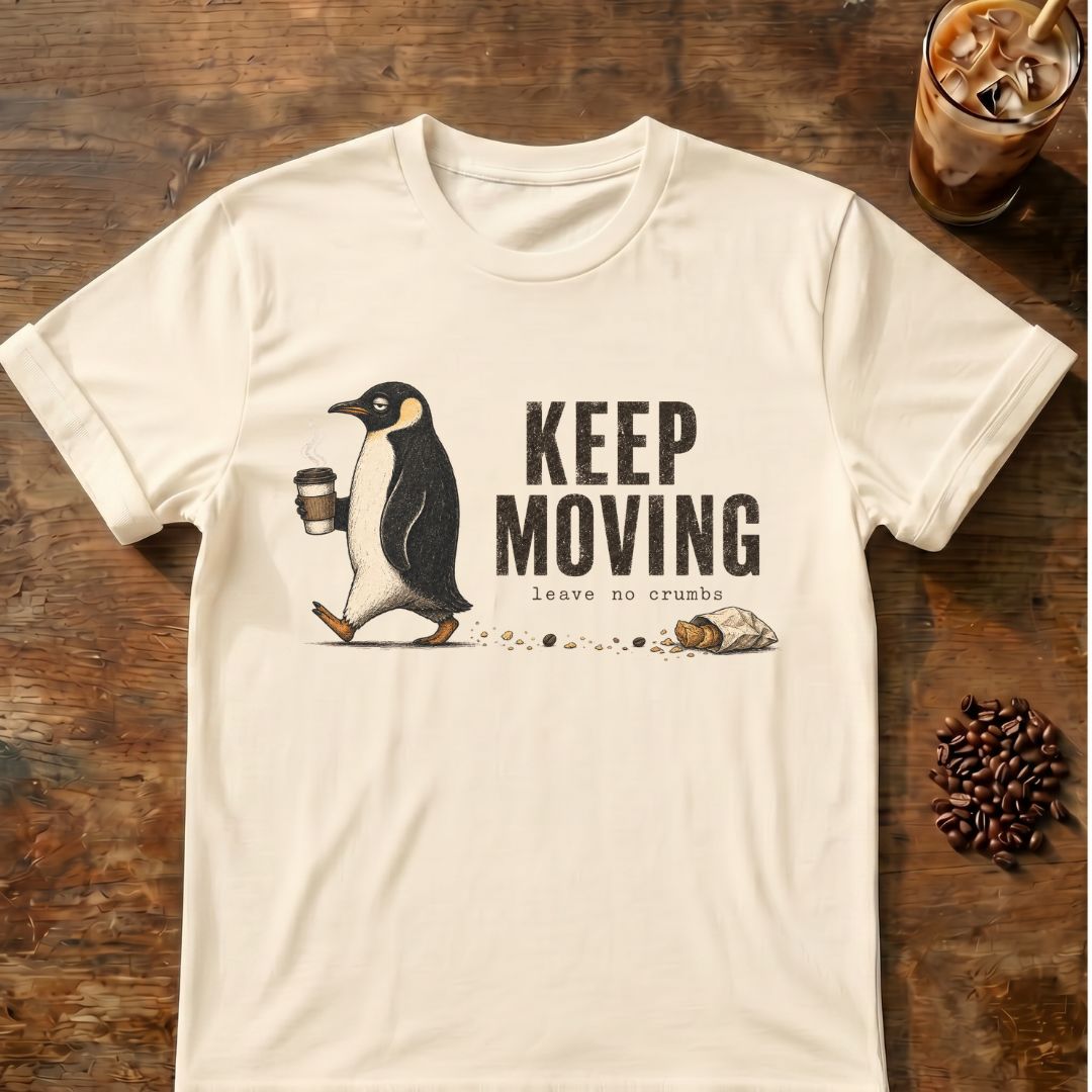 An off-white short-sleeve t-shirt with a graphic of a penguin walking and the text 'KEEP MOVING leave no crumb', displayed flat on a rustic wooden background with coffee-themed props.