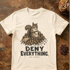An off-white short-sleeve graphic t-shirt laid flat on a rustic wooden table. The shirt features a detailed illustration of a squirrel sitting in a pile of coffee beans and holding a mug, with the text 'DENY EVERYTHING.