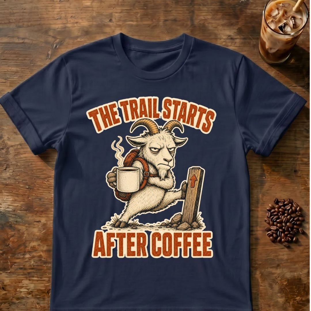 A navy blue graphic t-shirt laid flat on a rustic wooden surface, featuring a cartoon goat carrying a coffee mug with the text 'THE TRAIL STARTS AFTER COFFEE' in orange. Styled flat-lay props include an iced coffee and coffee beans.