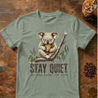 A sage green graphic t-shirt featuring a vintage-style illustration of a koala holding a mug on a branch, with the text 'STAY QUIET' and 'the mug knows too much'. The t-shirt is displayed flat on a rustic wooden table with coffee beans and an iced coffee in the background.