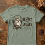 A sage green short-sleeve t-shirt with a graphic of an owl holding a coffee mug and the text 'RESPECT THE LOCALS SOME OF US WORK NIGHTS', displayed flat on a rustic wooden table with coffee beans and an iced coffee in the background.