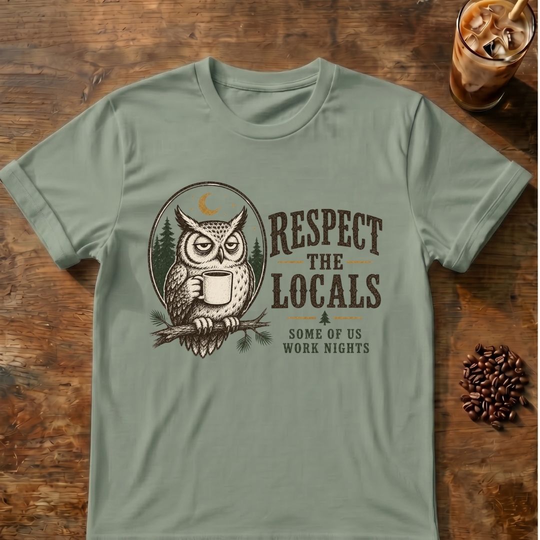 A sage green short-sleeve t-shirt with a graphic of an owl holding a coffee mug and the text 'RESPECT THE LOCALS SOME OF US WORK NIGHTS', displayed flat on a rustic wooden table with coffee beans and an iced coffee in the background.