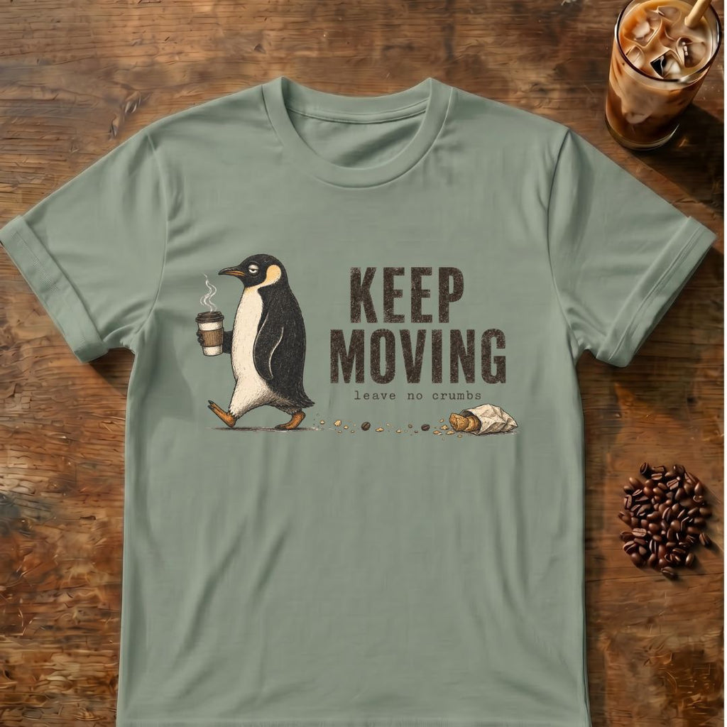 A sage green short-sleeve t-shirt with a graphic of a penguin walking and the text 'KEEP MOVING leave no crumb', displayed flat on a rustic wooden background with coffee-themed props