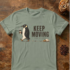 A sage green short-sleeve t-shirt with a graphic of a penguin walking and the text 'KEEP MOVING leave no crumb', displayed flat on a rustic wooden background with coffee-themed props