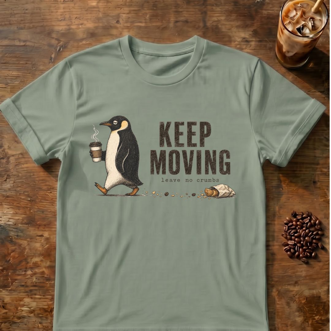 A sage green short-sleeve t-shirt with a graphic of a penguin walking and the text 'KEEP MOVING leave no crumb', displayed flat on a rustic wooden background with coffee-themed props