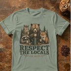 A sage green graphic t-shirt laid flat on a rustic wooden table, featuring an illustration of a bear, owl, raccoon, and fox sharing coffee mugs with the text 'RESPECT THE LOCALS' and 'EAT LIFE COFFEE LIFE'. A glass of iced coffee and coffee beans are styled as props in the corners.