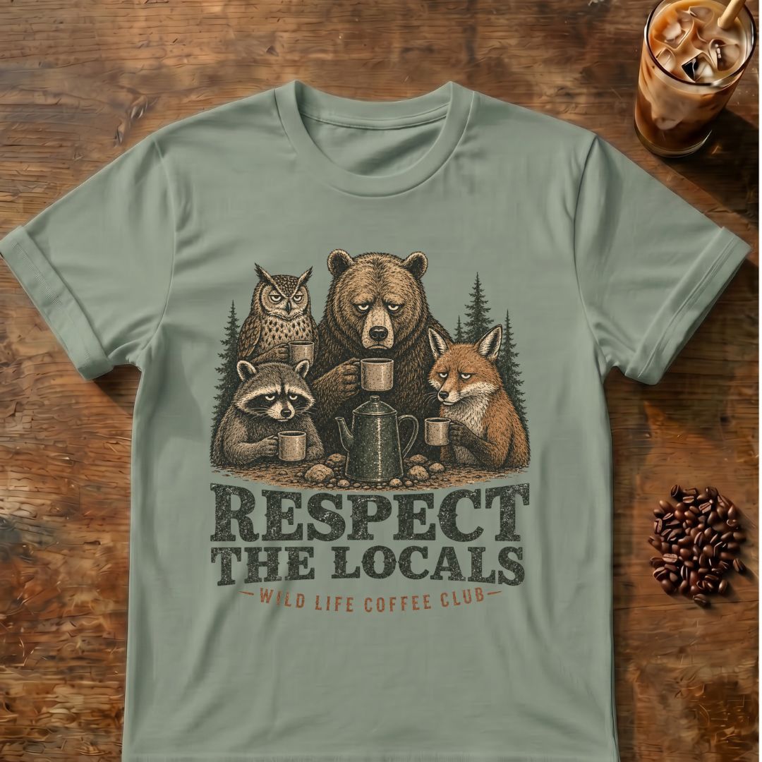 A sage green graphic t-shirt laid flat on a rustic wooden table, featuring an illustration of a bear, owl, raccoon, and fox sharing coffee mugs with the text 'RESPECT THE LOCALS' and 'EAT LIFE COFFEE LIFE'. A glass of iced coffee and coffee beans are styled as props in the corners.