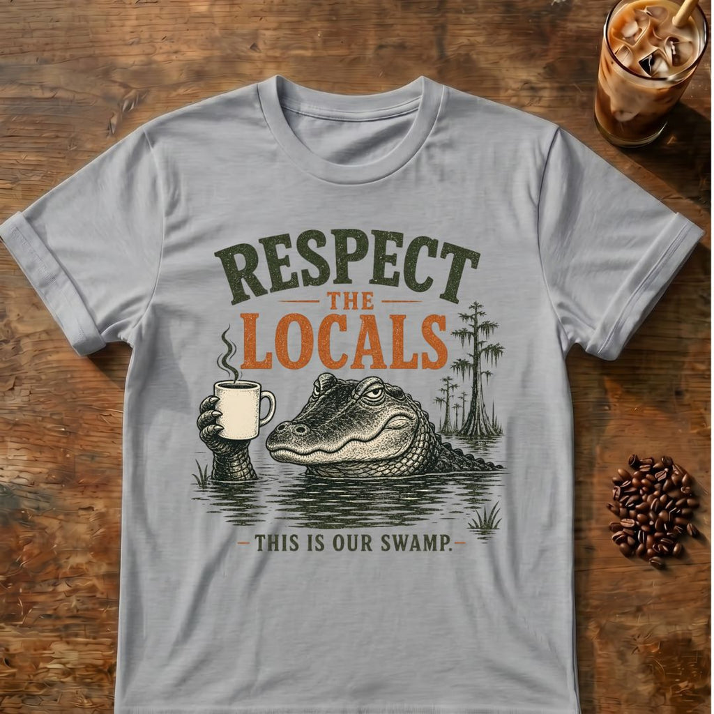 A light gray crewneck t-shirt laid flat on a rustic wooden table, featuring a graphic design of an alligator holding a mug with the text 'RESPECT THE LOCALS - THIS IS OUR SWAMP'. The shirt is styled with an iced coffee and coffee beans nearby.