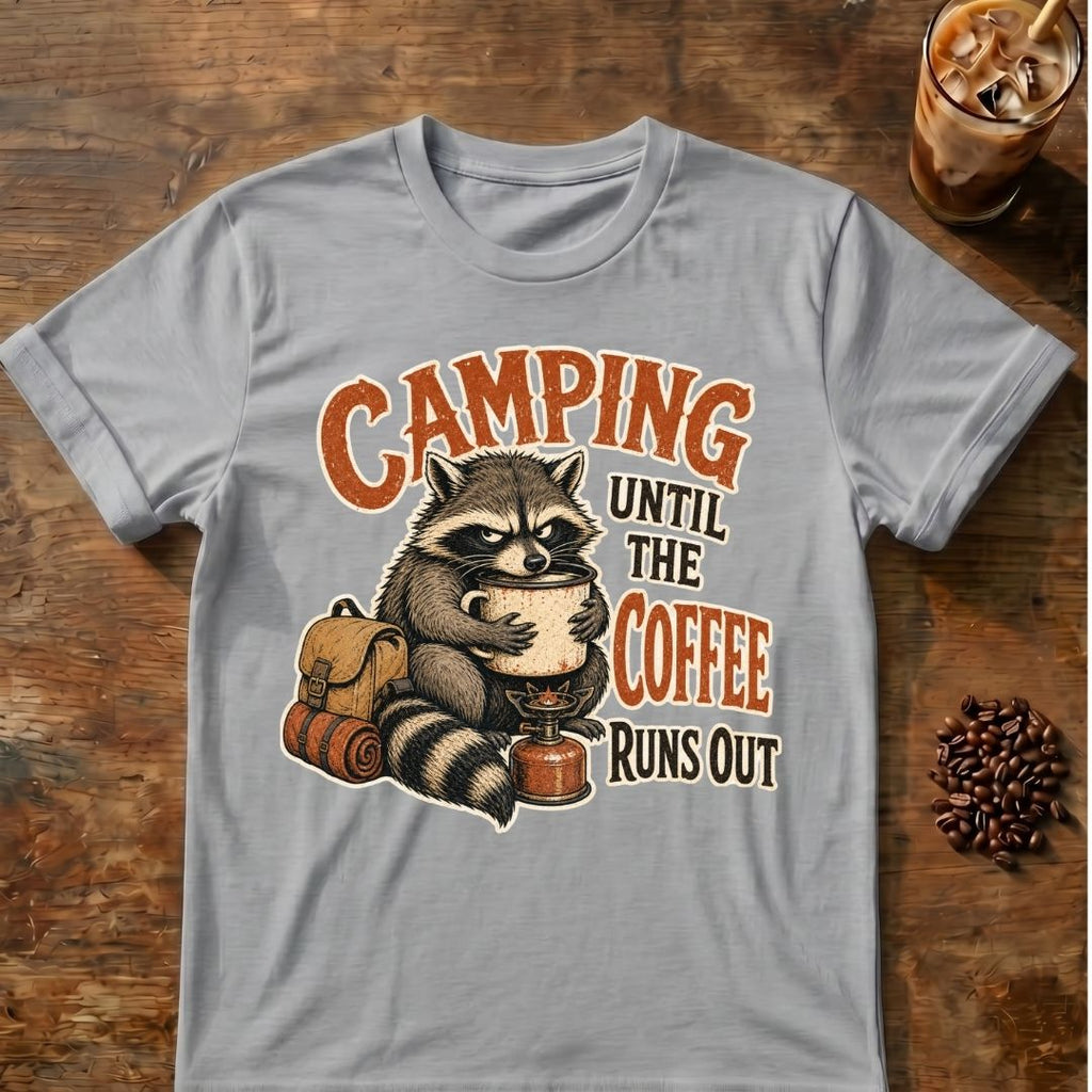 A light gray crewneck t-shirt featuring a graphic of a raccoon holding a coffee mug with a backpack and sleeping bag, with the text 'CAMPING UNTIL THE COFFEE RUNS OUT' in orange and brown, laid flat on a rustic wooden table with coffee props.