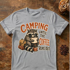 A light gray crewneck t-shirt featuring a graphic of a raccoon holding a coffee mug with a backpack and sleeping bag, with the text 'CAMPING UNTIL THE COFFEE RUNS OUT' in orange and brown, laid flat on a rustic wooden table with coffee props.