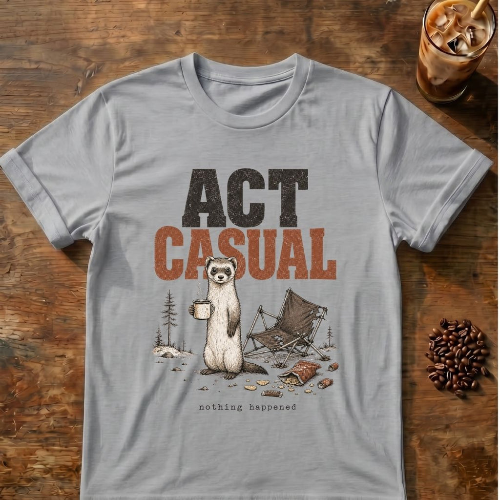 A light gray crewneck t-shirt with a graphic print of a standing ferret holding a mug next to a folding camp chair, with the text 'ACT CASUAL' above it. The shirt is displayed flat on a rustic wooden table with coffee beans and an iced coffee in the background.