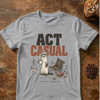 A light gray crewneck t-shirt with a graphic print of a standing ferret holding a mug next to a folding camp chair, with the text 'ACT CASUAL' above it. The shirt is displayed flat on a rustic wooden table with coffee beans and an iced coffee in the background.