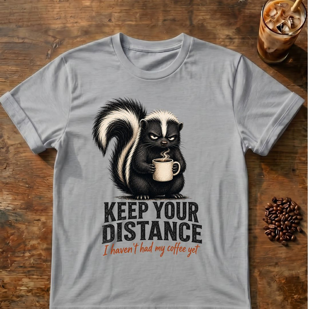 Grumpy skunk holding a steaming coffee mug above distressed text reading Keep Your Distance, with orange script saying I haven’t had my coffee yet. Sport Grey