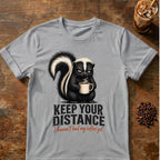 Grumpy skunk holding a steaming coffee mug above distressed text reading Keep Your Distance, with orange script saying I haven’t had my coffee yet. Sport Grey