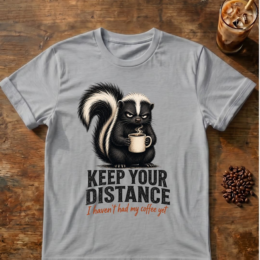 Grumpy skunk holding a steaming coffee mug above distressed text reading Keep Your Distance, with orange script saying I haven’t had my coffee yet. Sport Grey