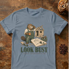 A light blue crewneck t-shirt featuring a cartoon beaver wearing green construction overalls and holding a coffee mug, standing next to a wooden shed with the text 'LOOK BUSY avoid responsibility' printed below. The t-shirt is displayed flat on a rustic wooden table with an iced coffee and coffee beans as props.