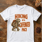 A white short-sleeve crewneck t-shirt laid flat on a rustic wooden table. The shirt features a graphic of a sloth hiking with a backpack and walking stick, next to the orange text 'HIKING AT A CAFFEINATED PACE'.