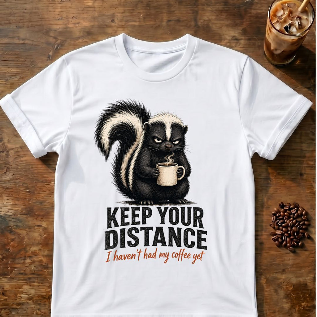 Grumpy skunk holding a steaming coffee mug above distressed text reading Keep Your Distance, with orange script saying I haven’t had my coffee yet. White