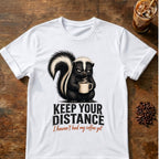 Grumpy skunk holding a steaming coffee mug above distressed text reading Keep Your Distance, with orange script saying I haven’t had my coffee yet. White