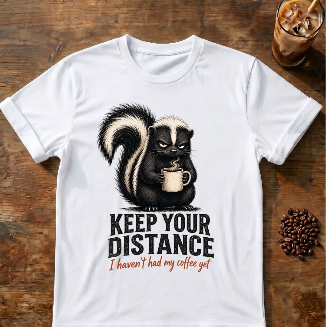 Grumpy skunk holding a steaming coffee mug above distressed text reading Keep Your Distance, with orange script saying I haven’t had my coffee yet. White