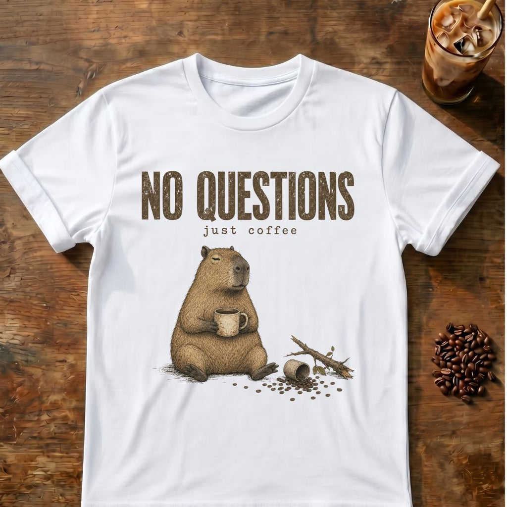 A white short-sleeve graphic T-shirt laid flat on a rustic wooden surface, featuring the text 'NO QUESTIONS just coffee' above an illustration of a capybara holding a mug with coffee beans and a broken branch nearby. A glass of iced coffee and scattered coffee beans appear as styled props around the shirt.